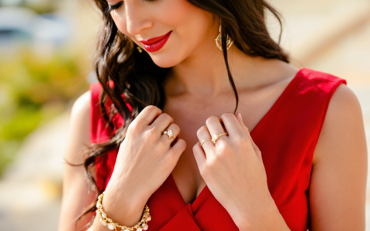 Une femme mettant en valeur une robe rouge avec des bijoux en or, se tenant dans un cadre extérieur lumineux, le soleil se reflétant sur les bijoux.