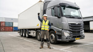 Conductrice de poids lourd en tenue de sécurité devant son camion sur une plateforme logistique française, photographiée en lumière naturelle dans un style reportage réaliste.
